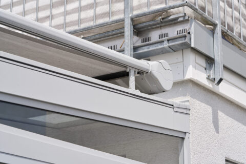 Graue Aufdachmarkise am Wintergarten, an Balkonbrüstung befestigt, Nahaufnahme des Motors und der Führungsschiene für optima…