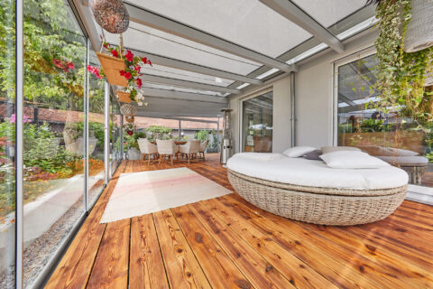 Moderner Ganzglas-Wintergarten mit integrierter Markise, Holzterrasse und komfortablen Outdoor-Möbeln, umgeben von grüner Na…