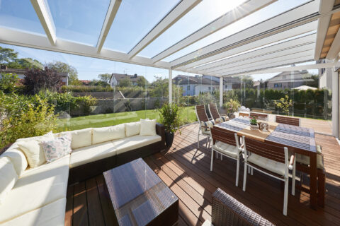 Moderner Kaltwintergarten aus Glas mit weißem Aluminiumrahmen, gemütlicher Sitzecke und Esstisch auf Holzterrasse mit Garten…