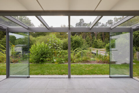 Moderner Kaltwintergarten mit transparenten Schiebeelementen, integriertem Sonnenschutz und Blick ins Grüne.