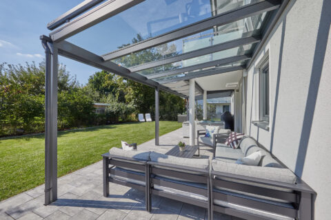 Moderne Glas-Terrassenüberdachung mit anthrazitfarbenem Aluminiumrahmen über einer stilvollen Outdoor-Lounge im Garten.