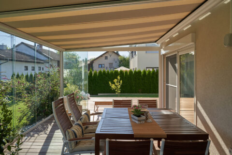 Moderner Kaltwintergarten mit Glaswänden, ausziehbarer Markise und Esstisch, der Blick in einen grünen Garten freigibt.
