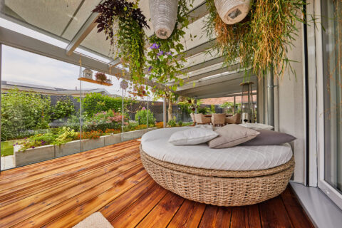 Moderner Glas-Wintergarten mit runder Lounge, Holzfußboden und Blick auf einen blühenden Garten – ideal für Entspannung.
