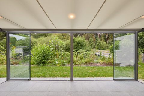 Blick aus einem modernen Kaltwintergarten mit Glasschiebetüren auf einen grünen Garten und Bäume. Heller Deckenbereich.